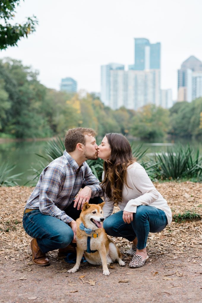couple kissing for their couple smiling and holding their dog for piedmont park engagement photos